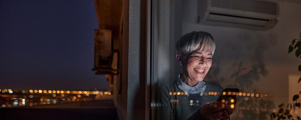 Glad mogen affärskvinna som använder mobiltelefon vid fönstret på natten.