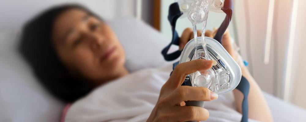 Person som ligger i en säng och håller i en CPAP-mask, en vanlig behandling vid sömnapné.
