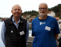Two men in their 70s are standing in front of the sea wearing shirts with "Kivik seaweed" printed on them and name tags.