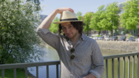 Studenten Taha står på en bro i Göteborg med högerhanden på huvudet där han har en hatt.